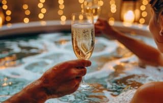 Man and woman ringing in the new year with a glass of champagne in a hot tub surrounded by holiday lights.